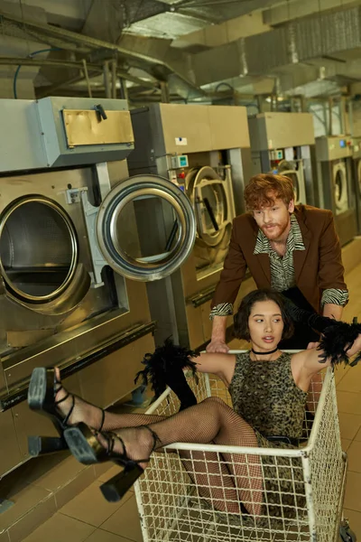 Uomo alla moda in piedi vicino asiatico fidanzata in carrello in pubblico lavanderia in sera — Foto stock
