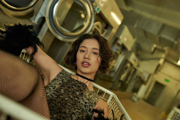 Trendige junge Asiatin in Kleid mit Animal Print sitzt im Einkaufswagen in der öffentlichen Wäscherei — Stockfoto