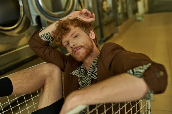 Jovem elegante ruiva homem no casaco olhando para a câmera enquanto sentado no carrinho na lavanderia moeda — Fotografia de Stock