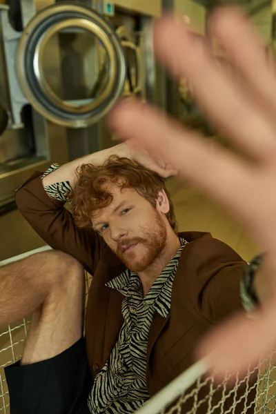 Young redhead man in jacket and trendy shirt looking at camera while sitting in cart in coin laundry — Stock Photo