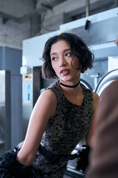 Стильна молода брюнетка азіатська жінка в одязі дивиться в сторону і позує в громадському пральні ввечері — стокове фото