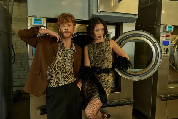 Joven pareja multiétnica posando en trajes elegantes cerca de la lavadora en la lavandería de monedas por la noche - foto de stock