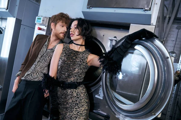 Trendy redhead man standing near asian girlfriend in dress ear washing machines in public laundry — Stock Photo