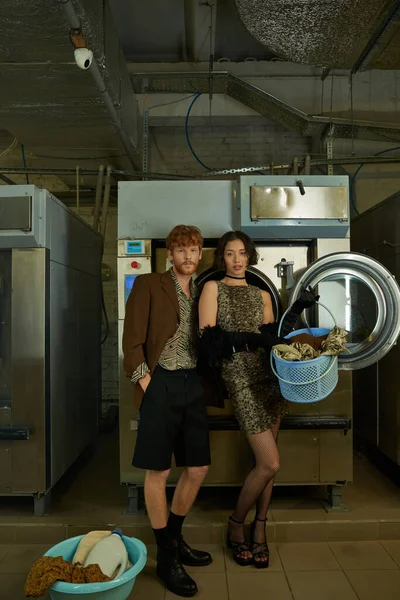 Casal interracial em roupas elegantes posando perto de roupas e máquinas de lavar roupa na lavanderia pública — Fotografia de Stock