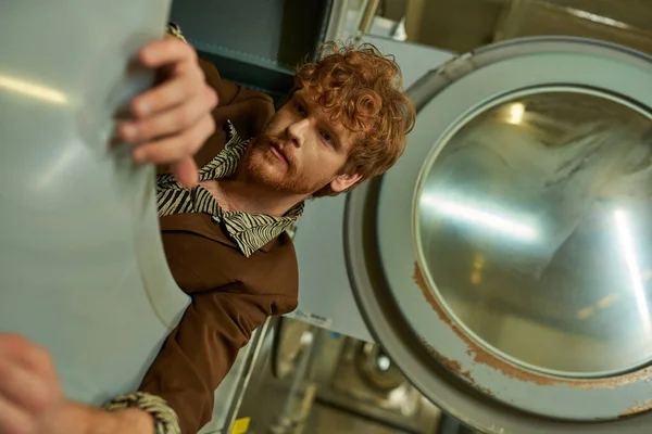 Fashionable young redhead man in jacket posing in washing machine in public laundry — Stock Photo