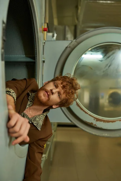 Стильный рыжий и бородатый молодой человек смотрит в камеру из стиральной машины в прачечной — стоковое фото