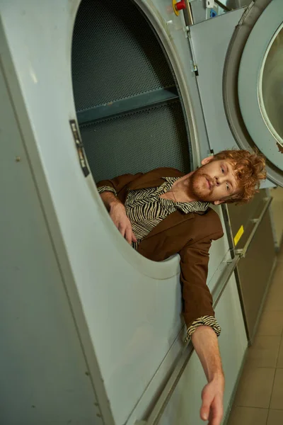 Молодой рыжий мужчина в современном наряде позирует в стиральной машине в прачечной самообслуживания — стоковое фото