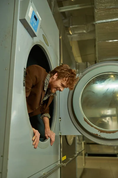 Fröhlicher und trendiger junger Rotschopf posiert in der Waschmaschine in der öffentlichen Wäscherei — Stockfoto