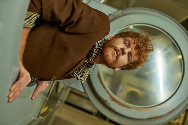 Portrait of young fashionable redhead man in jacket posing in washing machine in coin laundry — Stock Photo
