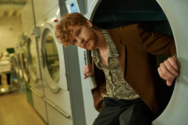Selbstbewusster junger Rotschopf blickt in die Kamera, während er in der Waschmaschine in der Münzwäsche posiert — Stockfoto