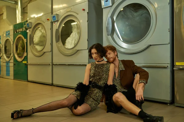 Moda casal interracial sentado perto de máquinas de lavar roupa na lavanderia moeda — Fotografia de Stock