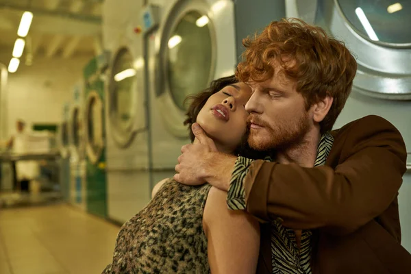 Sexy young man in stylish outfit touching asian girlfriend in dress in public laundry in evening — Stock Photo