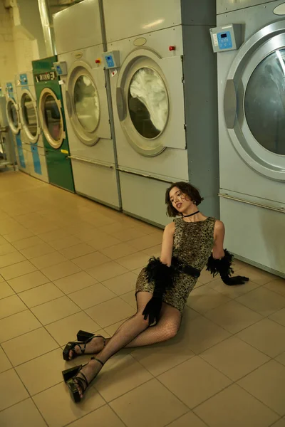 Sexy young asian woman in dress and gloves posing on floor in coin laundry in evening — Stock Photo