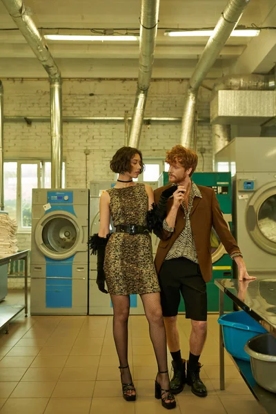 Fashionable interracial couple holding hands while standing together in coin laundry — Stock Photo