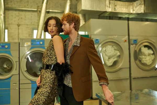 Sexy young man in jacket standing near asian girlfriend in dress in self service laundry — Stock Photo