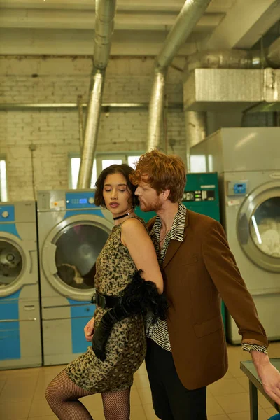 Jovem homem no casaco posando com elegante asiático namorada no vestido em moeda lavanderia — Fotografia de Stock