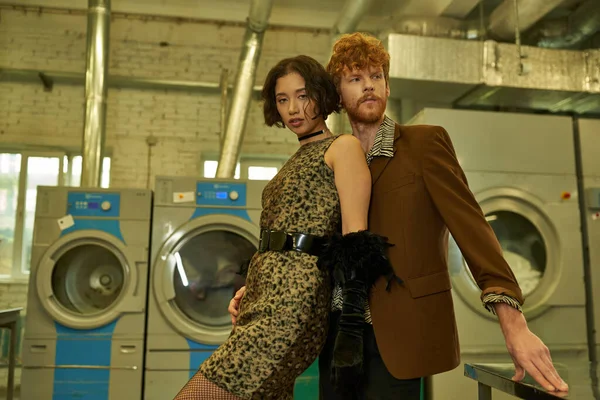 Fashionable asian woman in dress looking at camera near redhead boyfriend in self service laundry — Stock Photo