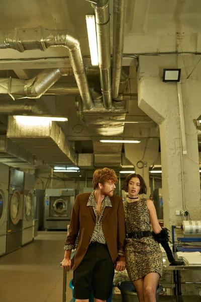 Young interracial couple in modern outfits posing while standing in public laundry in evening — Stock Photo