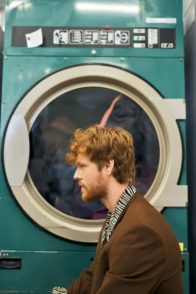 Seitenansicht eines modischen jungen rothaarigen Mannes, der in der Nähe einer Waschmaschine in der öffentlichen Wäscherei posiert — Stockfoto
