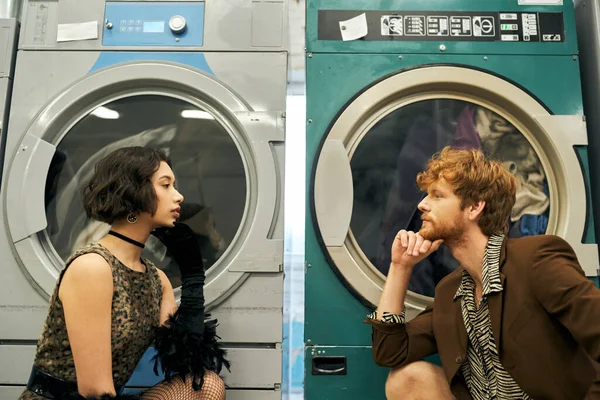 Vista lateral do moderno casal multiétnico olhando uns para os outros perto de máquinas de lavar roupa na lavanderia pública — Fotografia de Stock