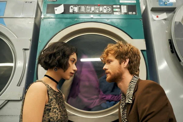Vista lateral do casal interracial romântico e elegante posando perto da máquina de lavar roupa na lavanderia moeda — Fotografia de Stock