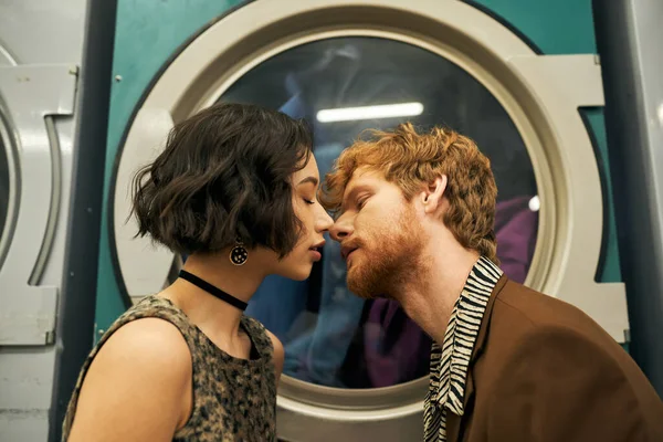 Jovem e elegante casal multiétnico beijando perto da máquina de lavar roupa na lavanderia moeda — Fotografia de Stock