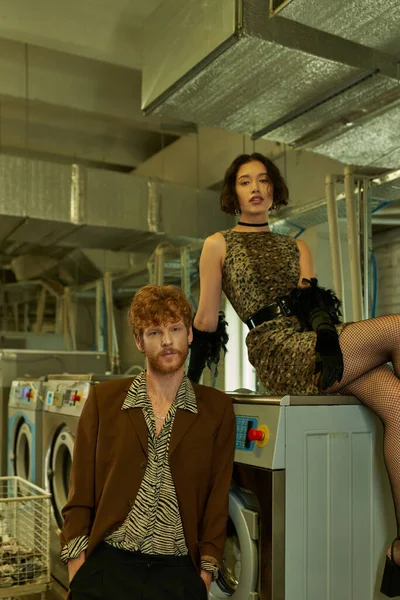 Fashionable young multiethnic couple in trendy outfits posing and looking at camera in coin laundry — Stock Photo