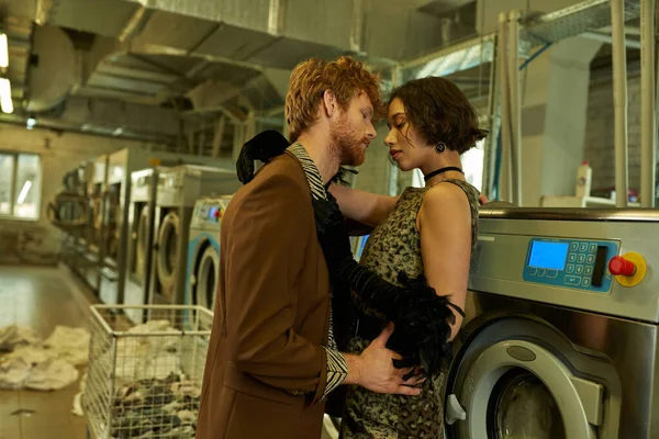 Стильная молодая межрасовая пара целуется возле стиральной машины в прачечной — стоковое фото