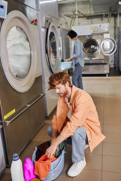 Jovem homem classificando roupas em cesta perto de detergentes e máquina de lavar roupa na lavanderia moeda — Fotografia de Stock