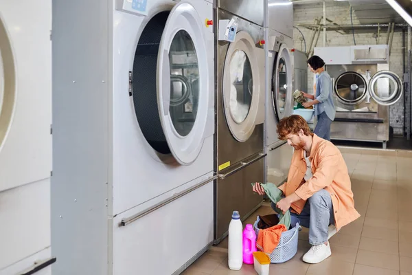 Young man taking clothes from basket near detergents and blurred girlfriend in public laundry — Stock Photo