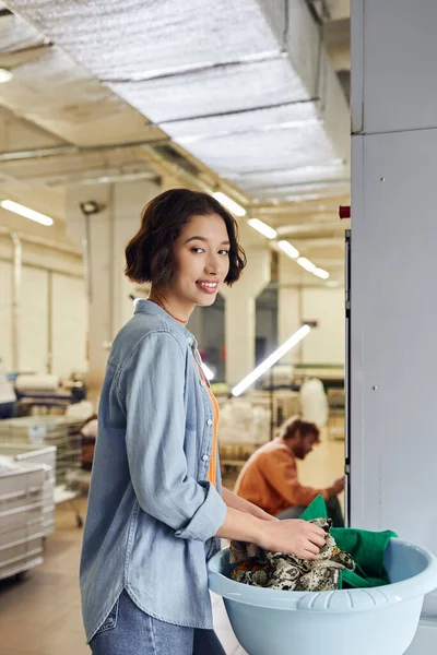 Smiling young asian woman looking at camera near clothes in basket in blurred public laundry — Stock Photo