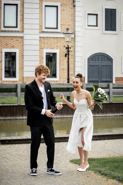 Young redhead groom opening champagne near african american bride with bouquet on street — Stock Photo