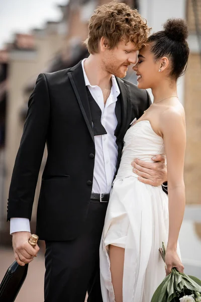 Young redhead man in elegant suit embracing happy african american bride, wedding outdoors — Stock Photo