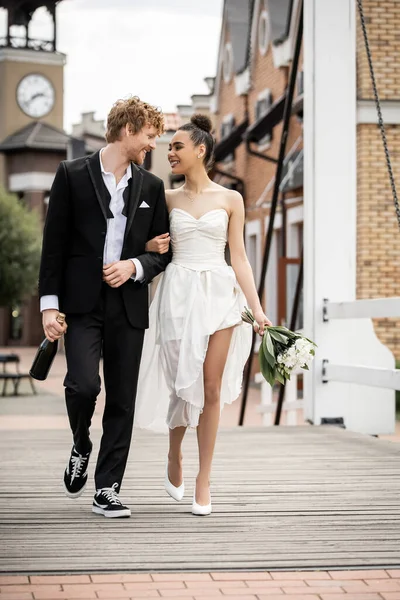 Boda al aire libre, multiétnicos recién casados con champán y flores caminando en el puente de la ciudad - foto de stock