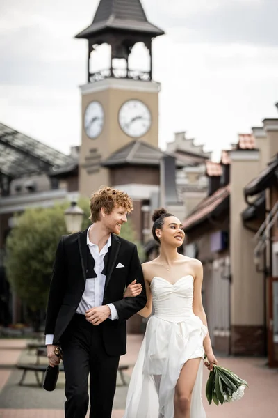 Celebración de la boda al aire libre, elegante y feliz pareja multiétnica caminando en la ciudad europea - foto de stock