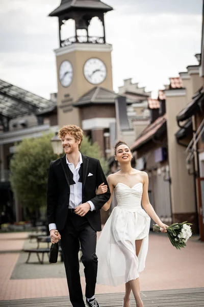 Romantic and cheerful interracial newlyweds with champagne and flowers walking on urban street — Stock Photo