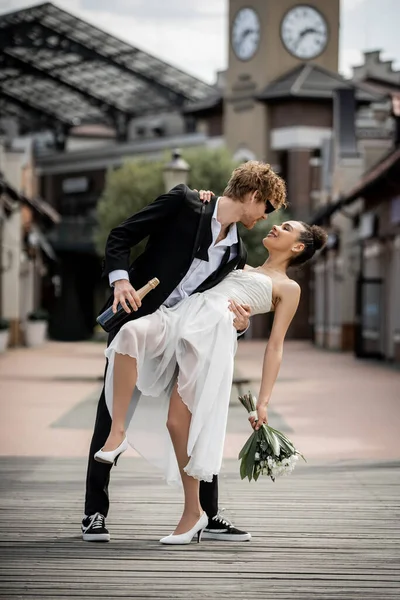 Celebración de la boda, ciudad europea, pelirroja novio abrazando elegante mujer afroamericana en la calle - foto de stock