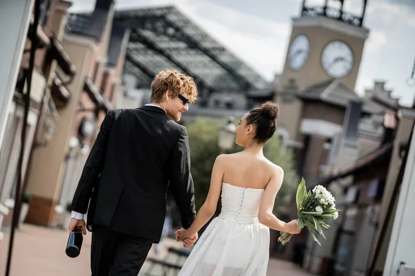 Urbane Romantik, Rückansicht multiethnischer Frischvermählter, mit Champagner, Blumen Händchen haltend auf der Straße — Stockfoto
