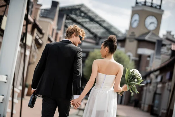Vue arrière de couple interracial élégant avec champagne et fleurs en ville, mariage en plein air — Photo de stock