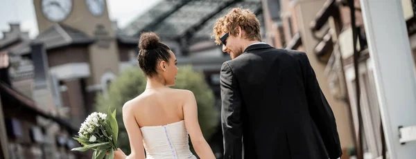 Back view of interracial newlyweds in elegant wedding attire on urban street, banner — Stock Photo