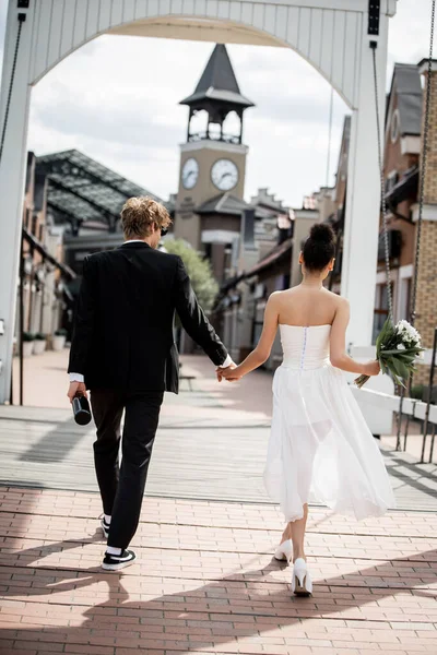 Vista trasera de pareja multiétnica con champán y flores cogidas de la mano en el puente, boda al aire libre - foto de stock