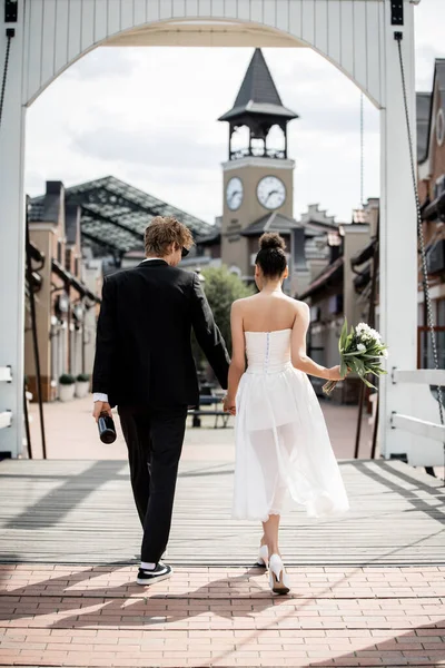 Back view of romantic multiethnic couple holding hands and walking on bridge, outdoors wedding — Stock Photo