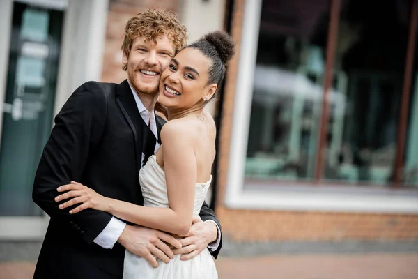 Joyeux et élégant multiethnique jeunes mariés étreignant et regardant la caméra dans la rue, mariage à l'extérieur — Photo de stock