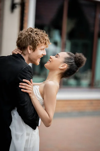 Vue latérale du jeune marié embrassant la mariée afro-américaine ravie en ville, célébration en plein air — Photo de stock