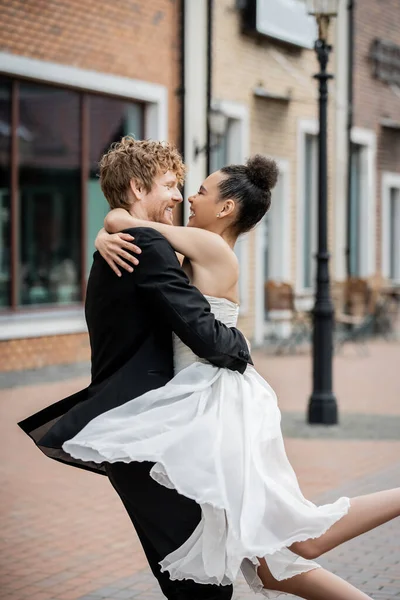 Pareja interracial en traje de boda abrazando en la calle de la ciudad, felicidad, boda al aire libre, vista lateral - foto de stock