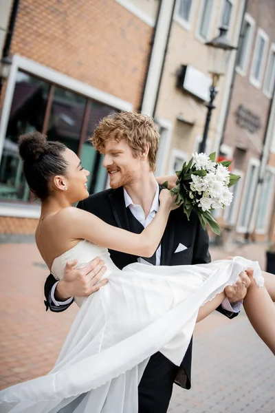 Romance urbaine, marié rousse tenant mariée afro-américaine avec bouquet de mariage dans la rue — Photo de stock