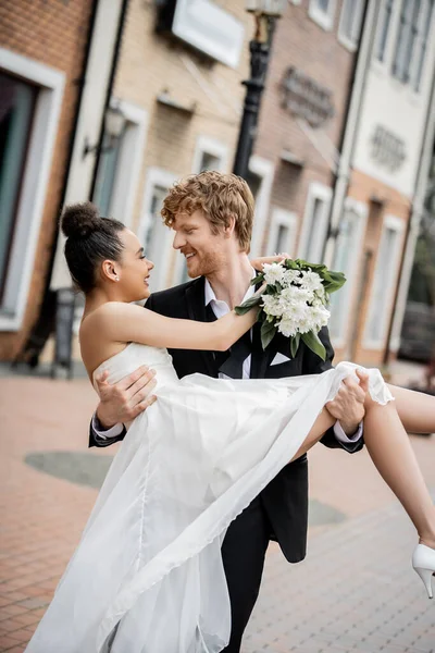 Amour en ville, jeune marié heureux tenant mariée afro-américaine avec des fleurs dans la rue — Photo de stock
