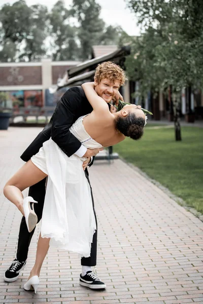 Homme souriant et femme afro-américaine en tenue de mariage embrassant dans la rue, célébration en plein air — Photo de stock