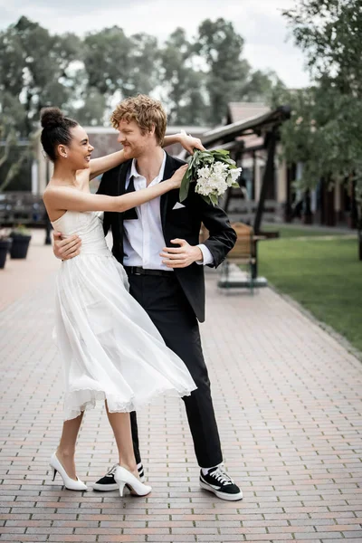Elegante afrikanisch-amerikanische Braut mit Strauß umarmt lächelnden Bräutigam auf der Straße, Freude, urbane Romantik — Stockfoto