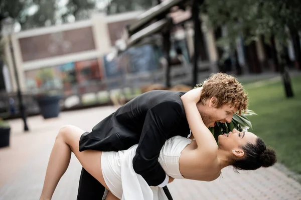 Jeune homme excité embrassant élégante mariée afro-américaine sur la rue de la ville européenne, vue de côté — Photo de stock
